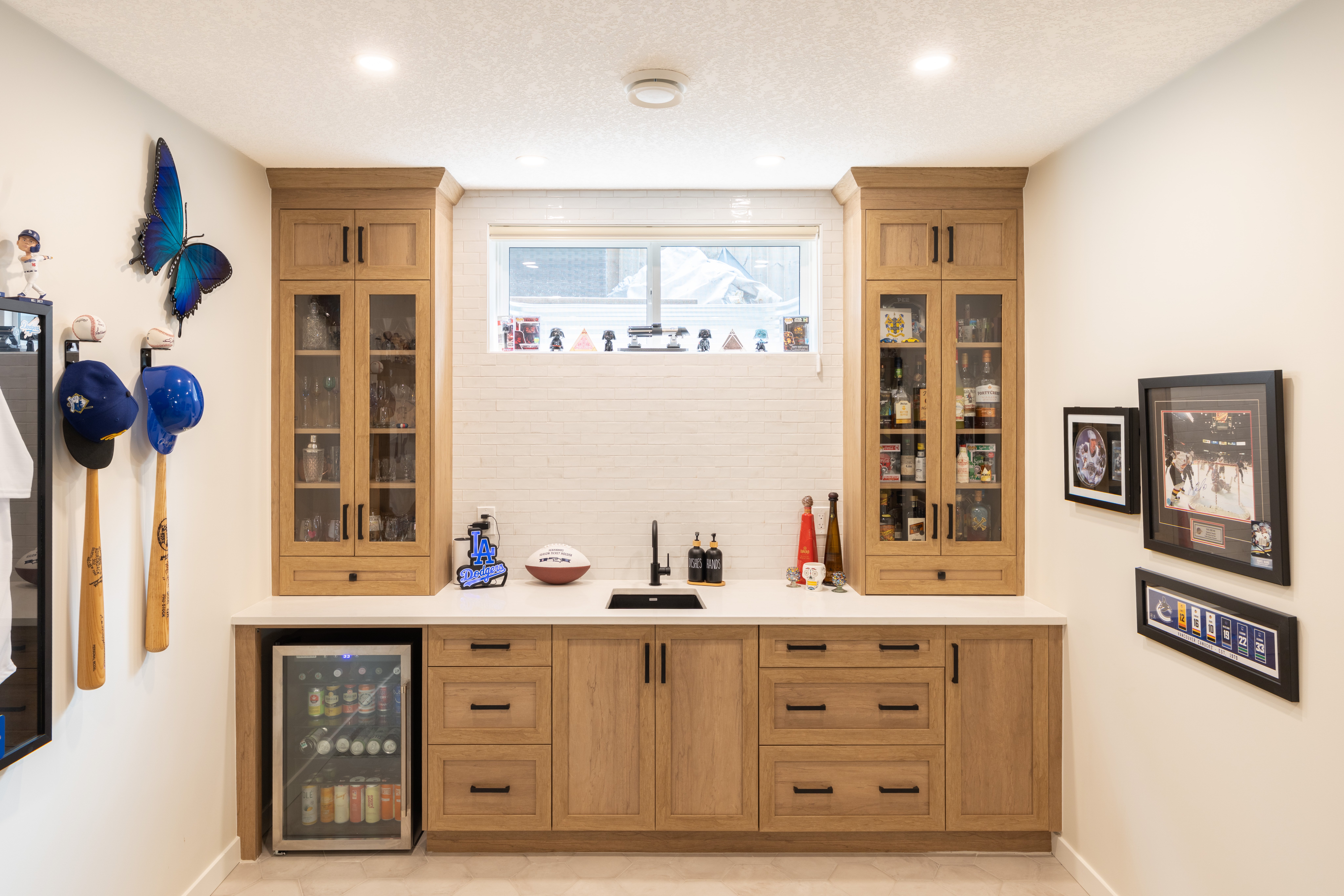Shaker-style cabinetry, glass-front display cabinets, a white countertop, and an undercounter beverage fridge complete the basement wet bar