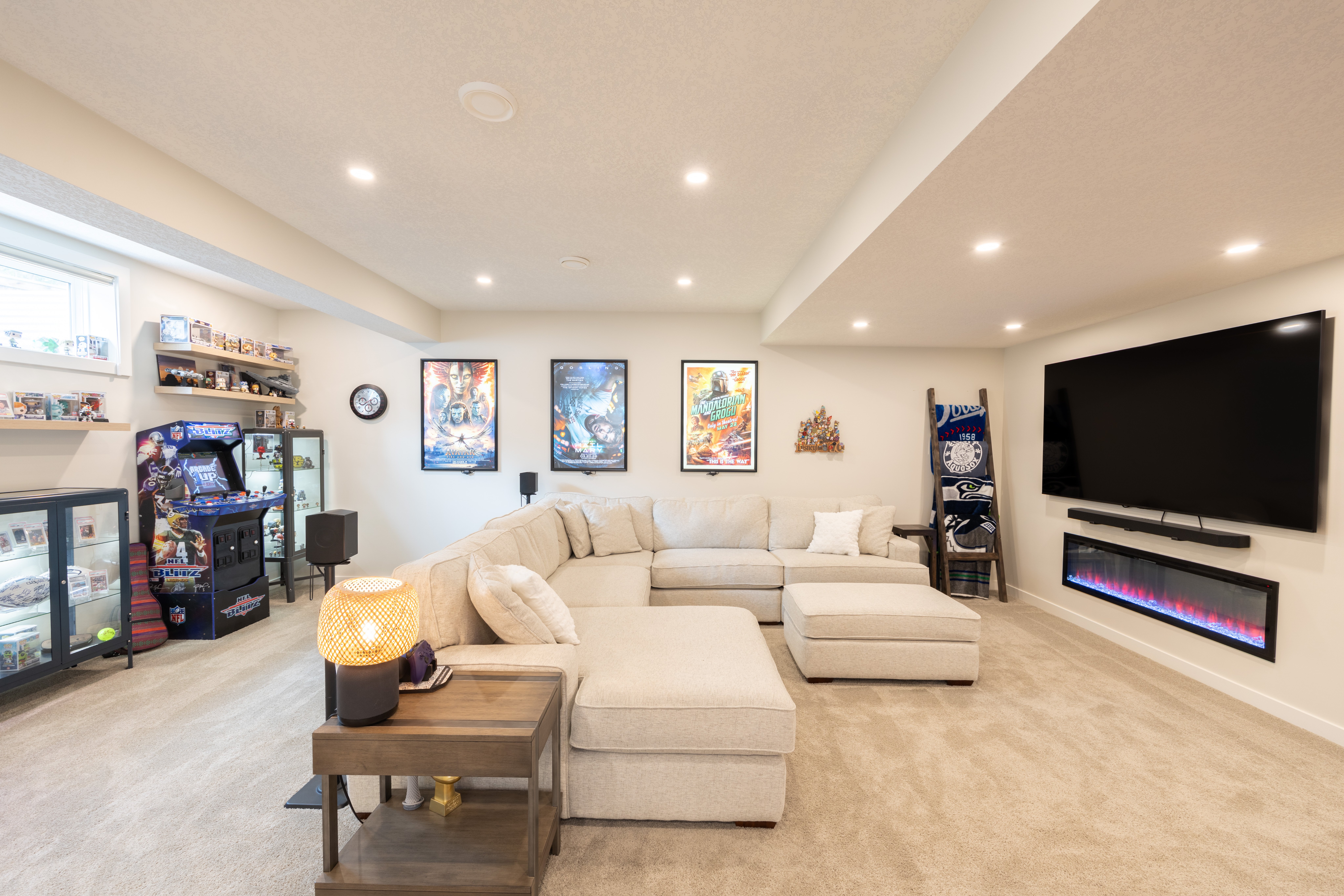 A spacious basement lounge with a pale sectional, wall-mounted television, recessed electric fireplace, and adjoining arcade area in Mahogany, Calgary.