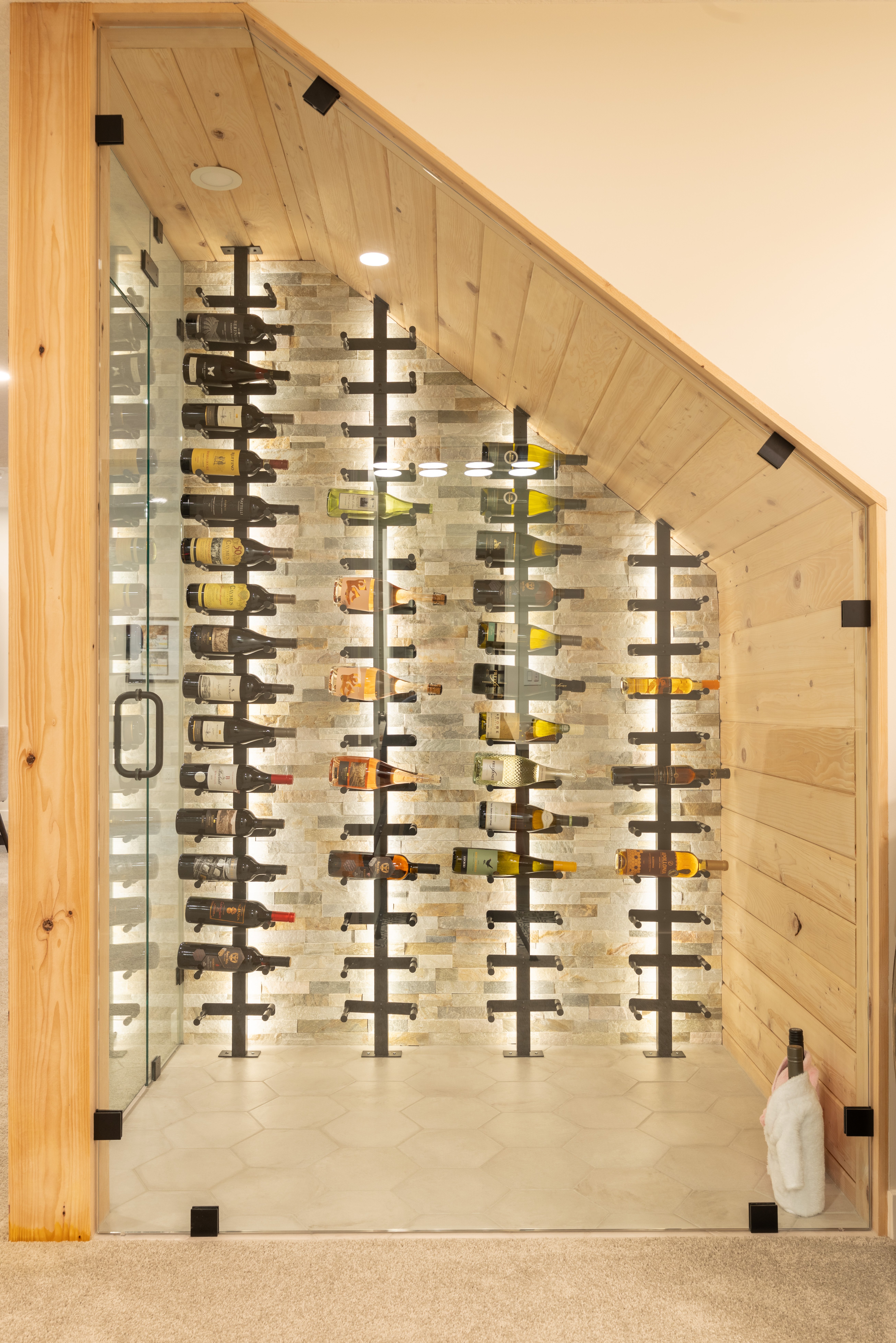 Black bottle racks stand against illuminated textured cladding inside a glass enclosure with wood lining and hexagonal floor tile.