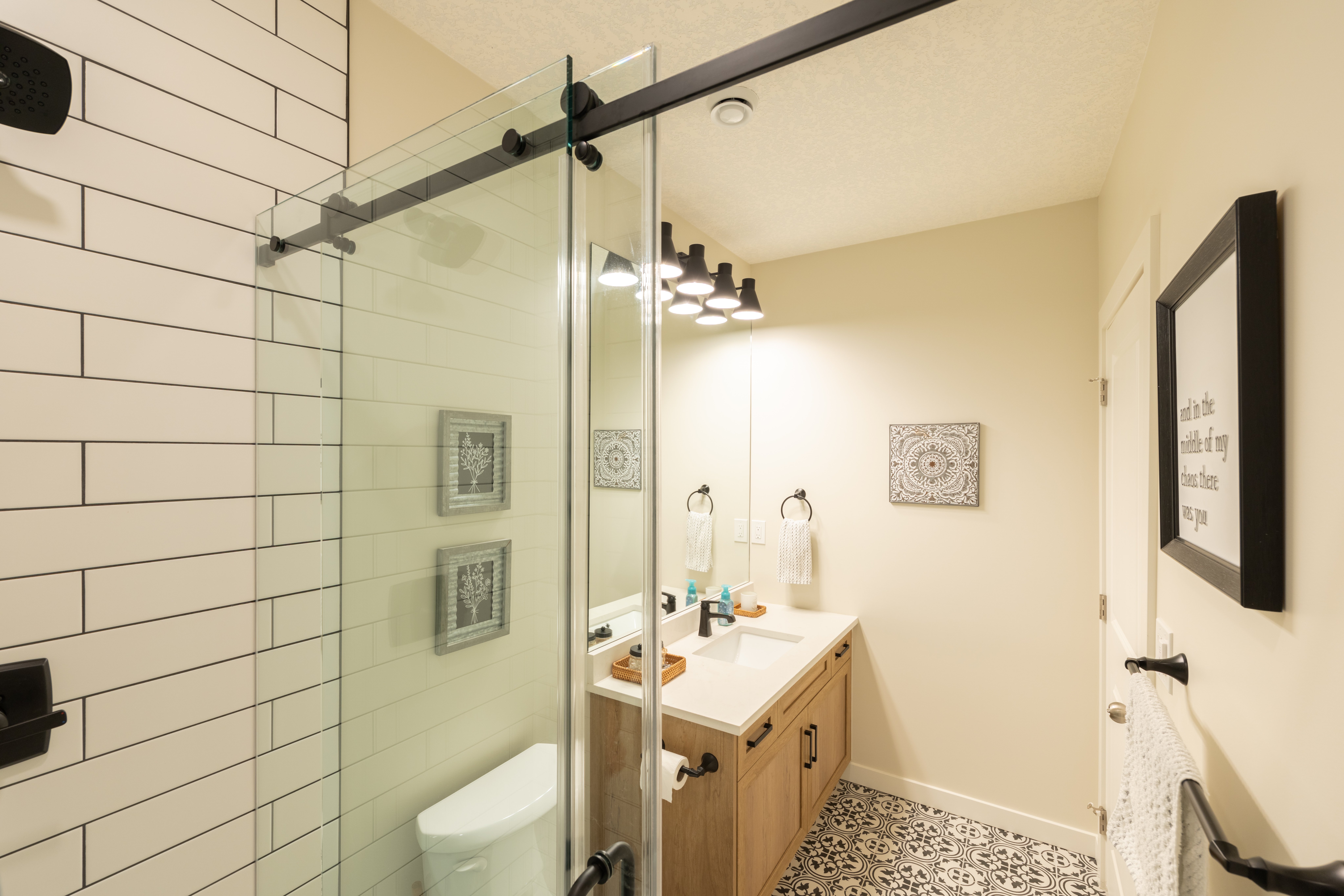 The shower’s clear glass panels and black sliding rail frame a view toward the bathroom vanity and patterned floor.