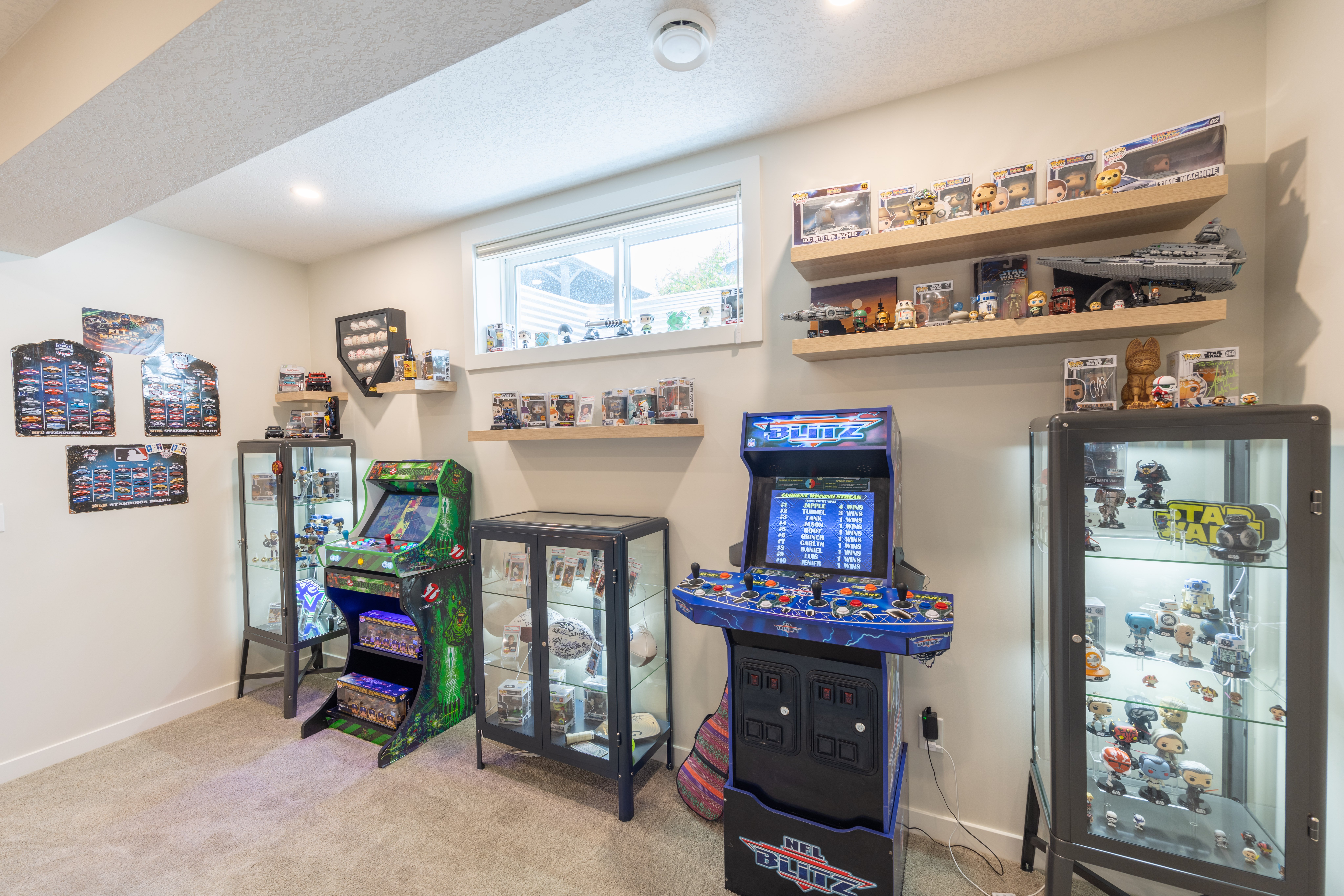 Arcade games, illuminated glass cabinets, and floating shelves create a dedicated display and gaming area beneath a basement window.